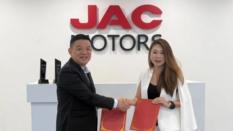 Lau Yit Mun and Connie Go exchanging signed document folders and shaking hands in front of a white wall featuring a large red JAC Motors logo.