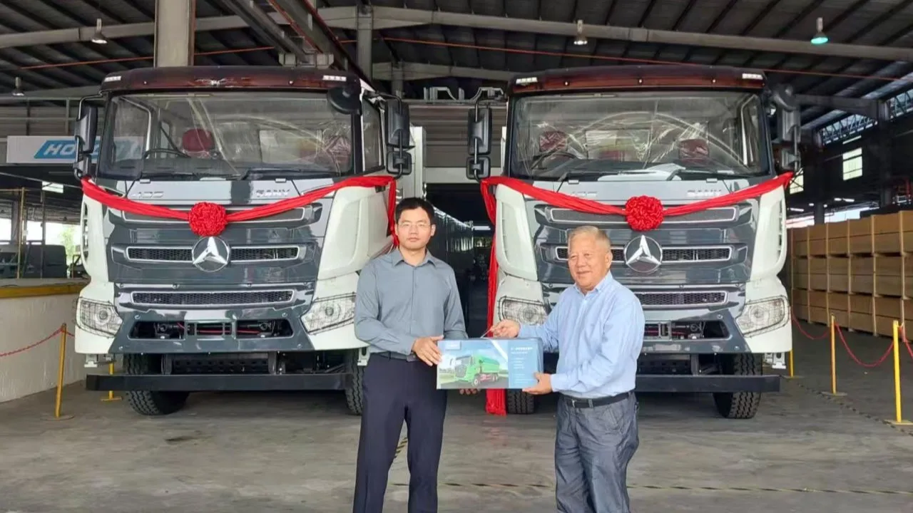 SANY Regional Chairman Liao Wei handing a boxed gift to Gemilang Coachwork Founder Pang Chong Yong while standing in front of two grey and white SANY dump trucks adorned with red ceremonial bows.