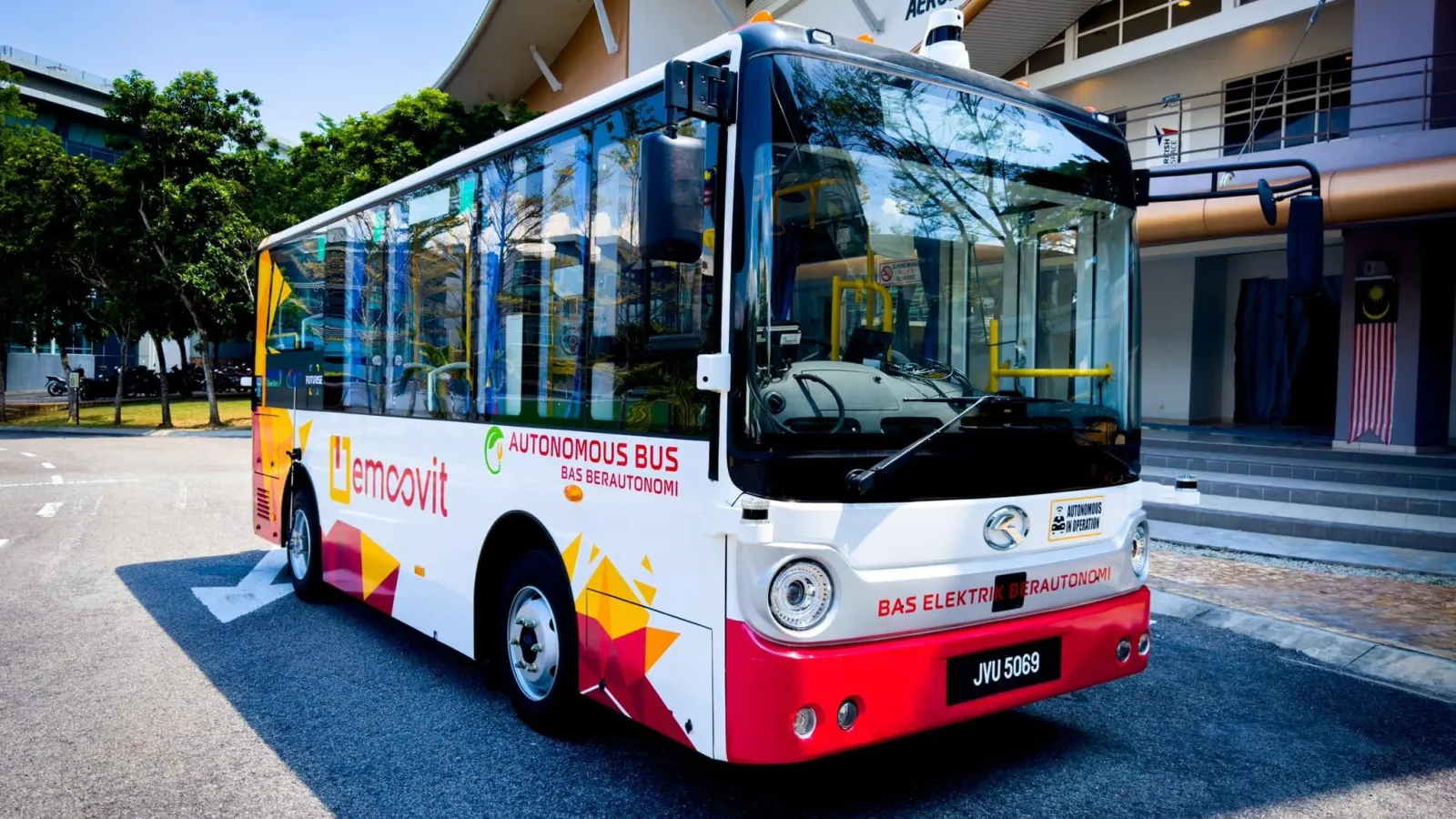 A compact white, red, and yellow autonomous electric bus with "eMoovit" and "Bas Berautonomi" decals parked in front of a modern civic building.