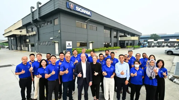 Dr Michael Jackstein, Carolin Riethus, and Scania Southeast Asia staff wearing blue shirts giving thumbs-up gestures outdoors in front of the Scania Bukit Jelutong headquarters building.