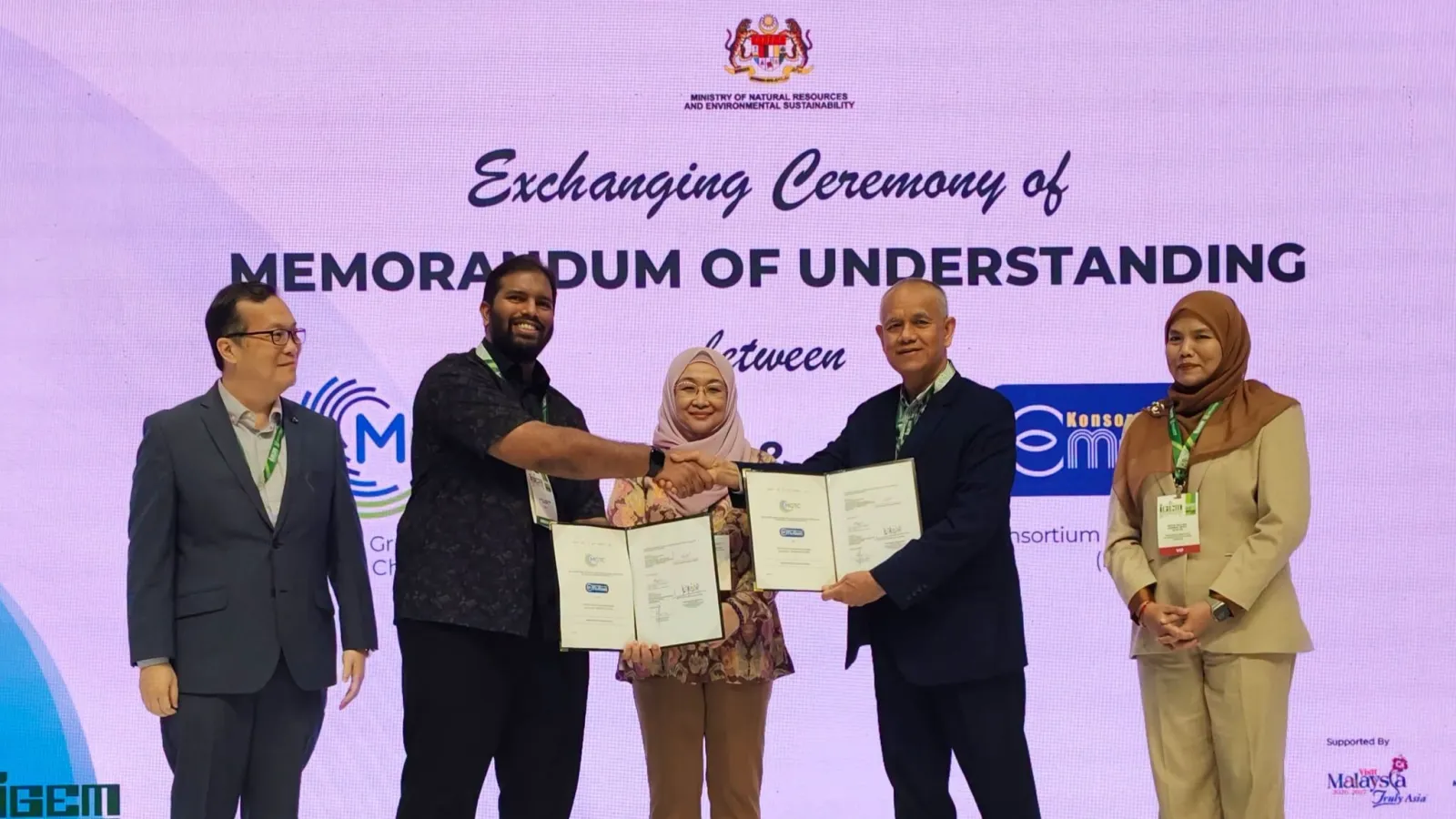 Representatives from MGTC and Konsortium E-Mutiara holding open document folders during a formal MoU exchange ceremony on stage in front of a Ministry backdrop.