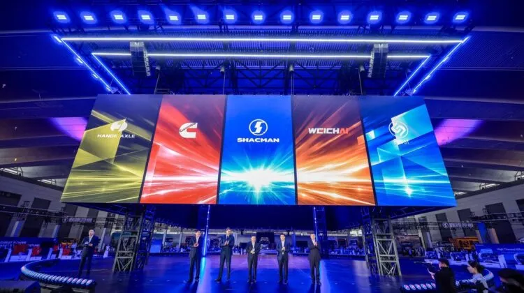 Five executives in business suits standing on a stage illuminated by blue lights in front of a giant digital screen displaying the logos of HanDe Axle, Cummins, Shacman, Weichai, and Fast.
