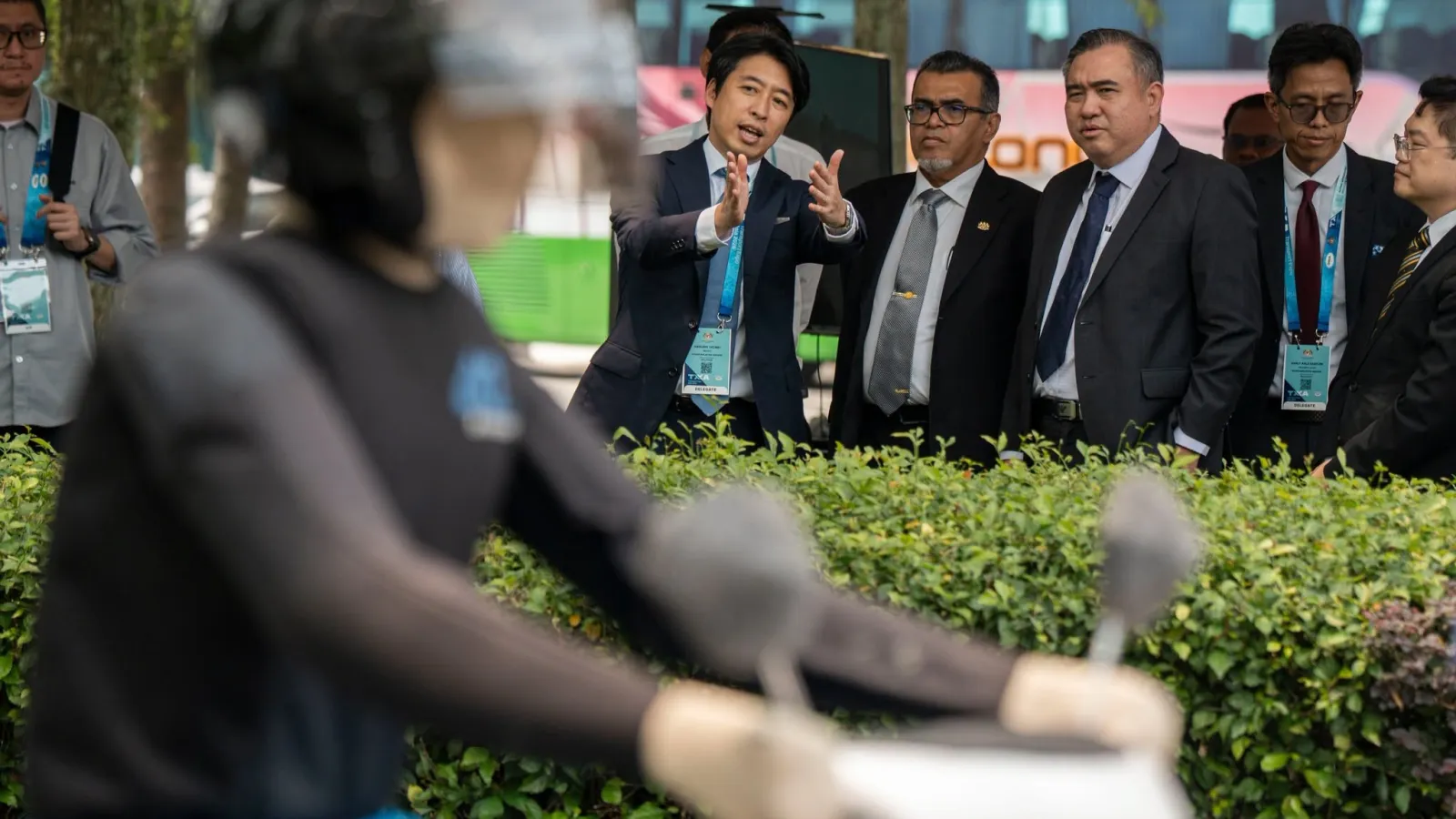 Transport Minister Anthony Loke in a suit watching an outdoor live demonstration of a test dummy riding a motorcycle behind green hedges.