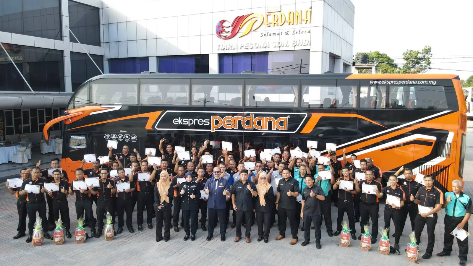 A group photo of express bus drivers in uniform holding completion certificates posing alongside JPJ officers and corporate executives in front of a black and orange Ekspres Perdana double-decker bus.