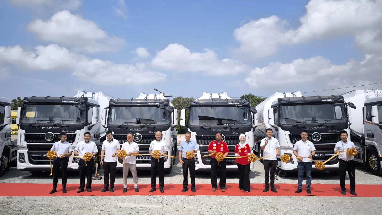 A lineup of company executives and guests holding a golden ribbon across a red carpet in front of four white Shacman X5000 Pro prime mover trucks parked outdoors.