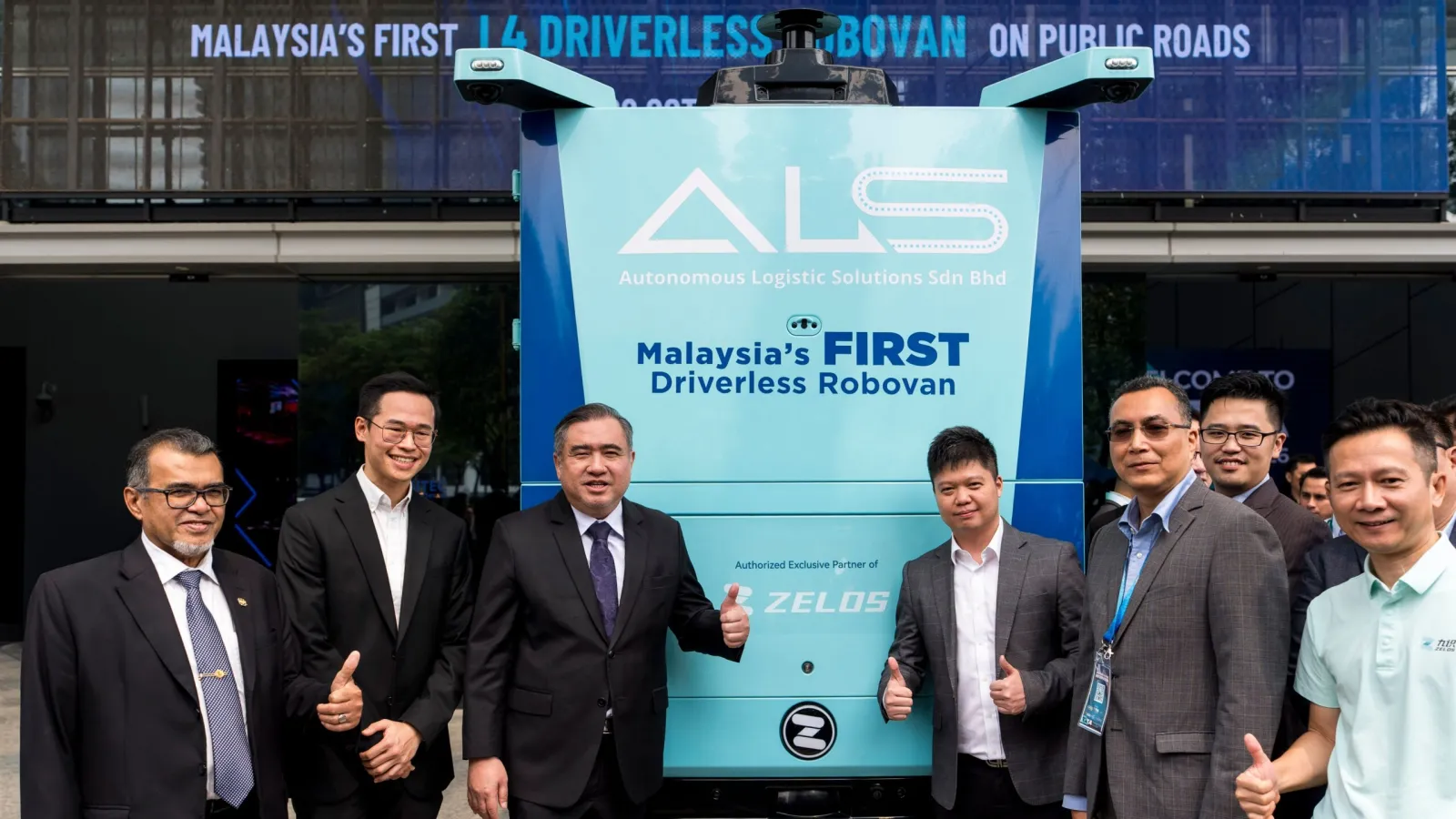 Transport Minister Anthony Loke giving a thumbs-up next to executives standing in front of a teal and white Zelostech Autonomous Logistic Solutions RoboVan.