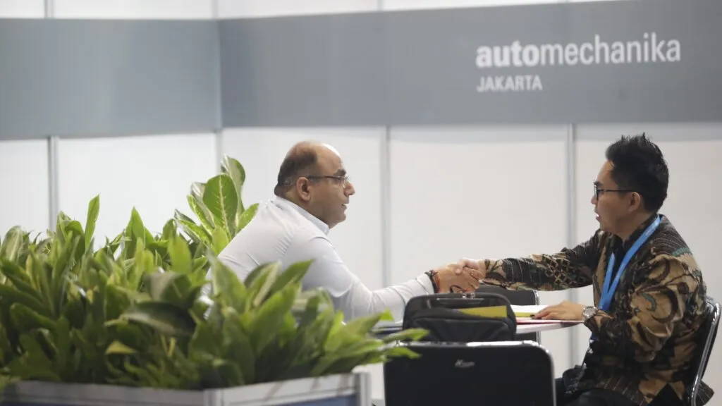 Two men in business attire shaking hands across a meeting table in front of potted green plants and a gray booth wall bearing the Automechanika Jakarta logo.