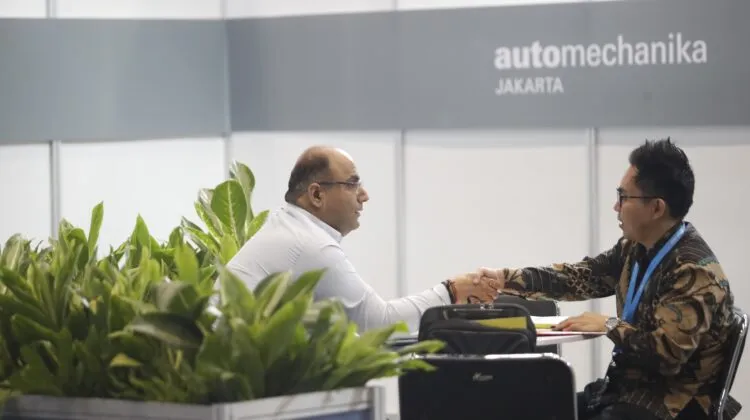 Two men in business attire shaking hands across a meeting table in front of potted green plants and a gray booth wall bearing the Automechanika Jakarta logo.